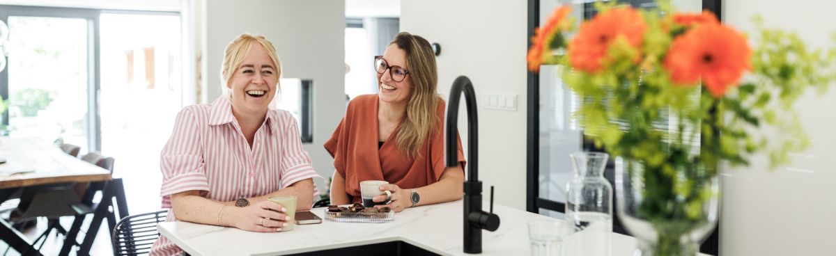 Twee lachende vrouwen met koffie in een gezellige setting aan het keukenblok in huis